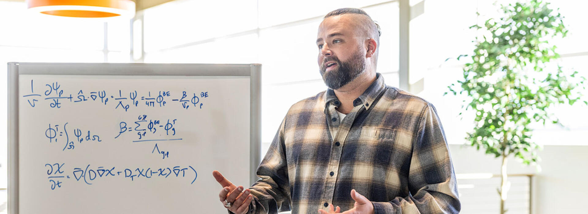 A person wears a plaid shirt and stands indoors near a whiteboard with complex mathematical equations.