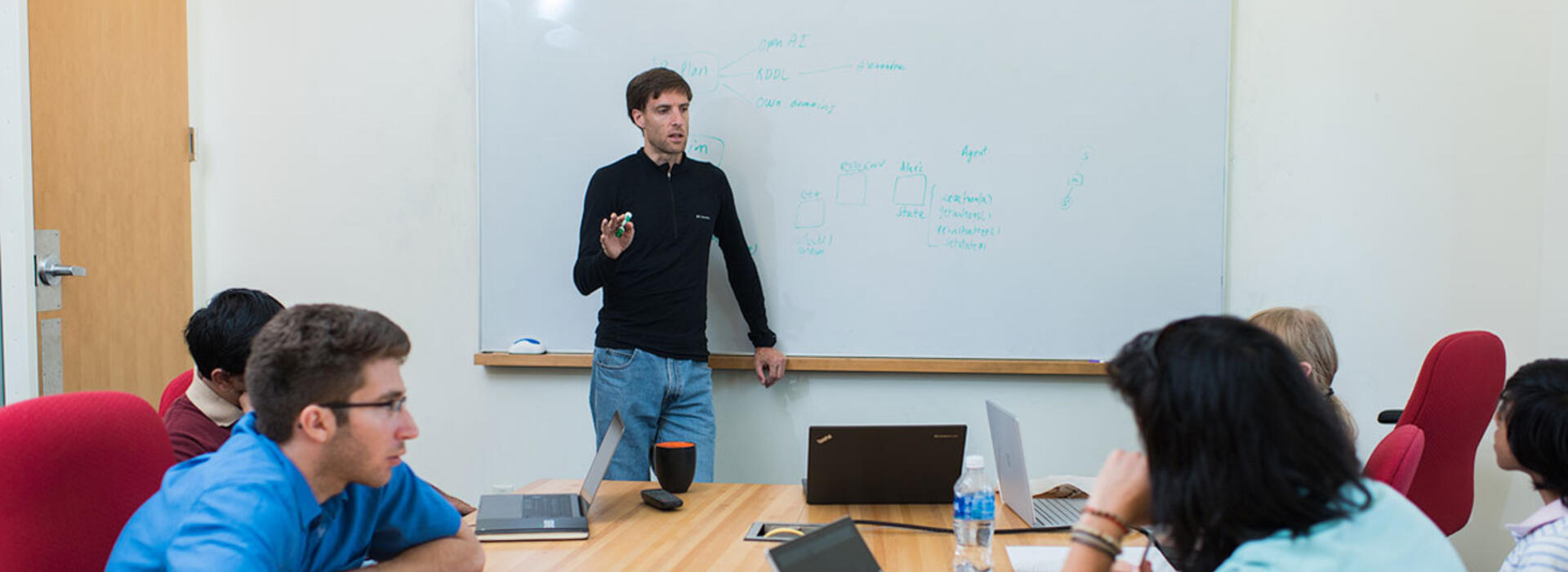 A person stands at the front of a conference room, pointing to a whiteboard covered with handwritten diagrams and notes, while several people sit around a table with laptops and notebooks, listening and discussing.