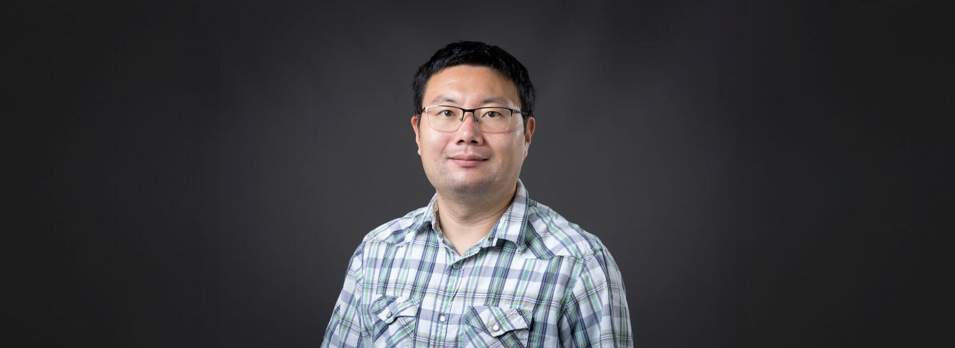 A person wears a light plaid button-up shirt and stands against a plain dark background, facing the camera.