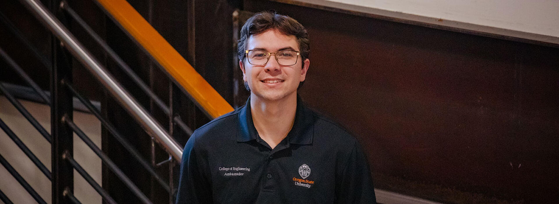 A person wears a black Oregon State University College of Engineering Ambassador polo shirt and stands indoors near a staircase with metal railings and dark wood paneling in the background.