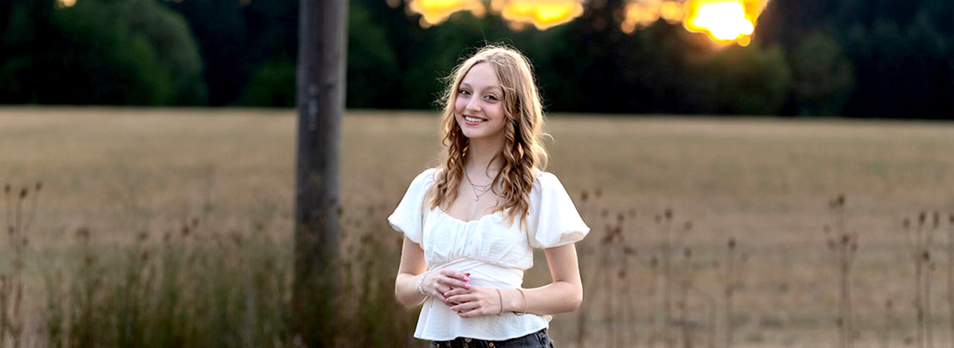 A person with shoulder-length hair stands in a field at sunset and wears a white short-sleeve shirt. 