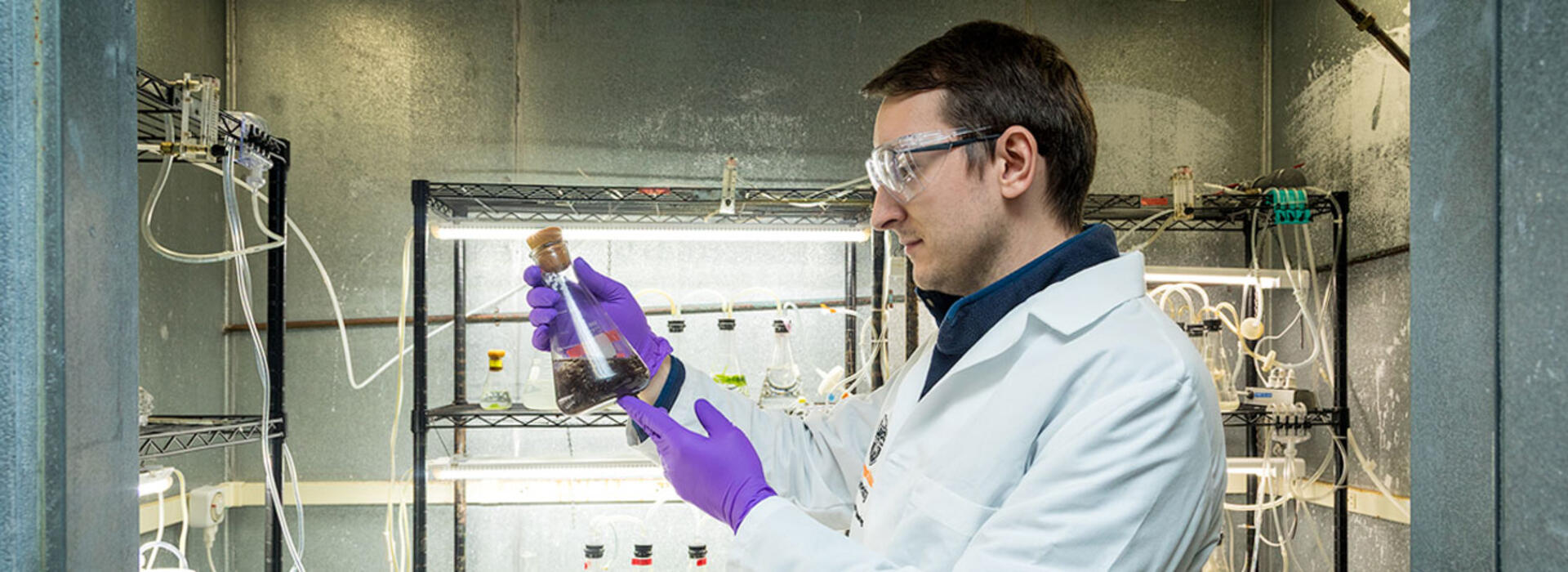 A person in a lab wears a lab coat and gloves while holding and examining a glass flask containing a dark liquid, standing in front of metal shelving.