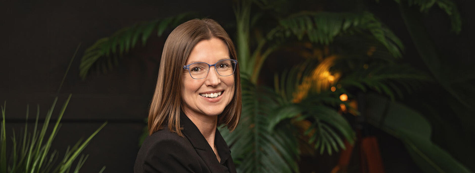 Camille Palmer wears business attire and sits in a dark area with tropical vegetation in the background and smiles toward the camera. 