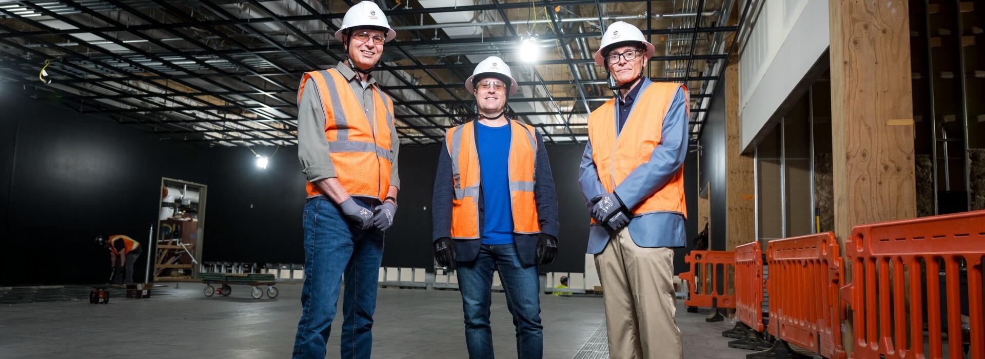 Anthony Koppers, Shayne Huddleston, and Todd Palmer dressed in hard hats and safety vests in a dark room under construction.
