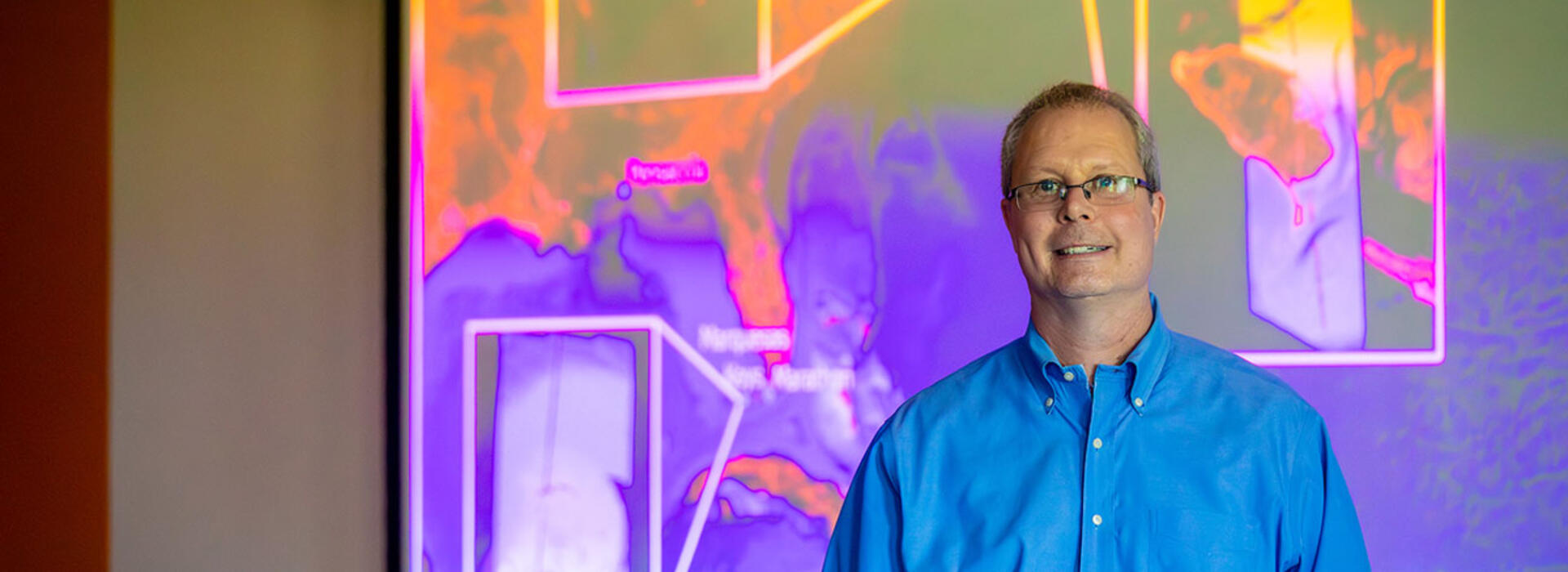 A person in business attire stands in a classroom with a digital projection of colorful bathymetry images behind them.