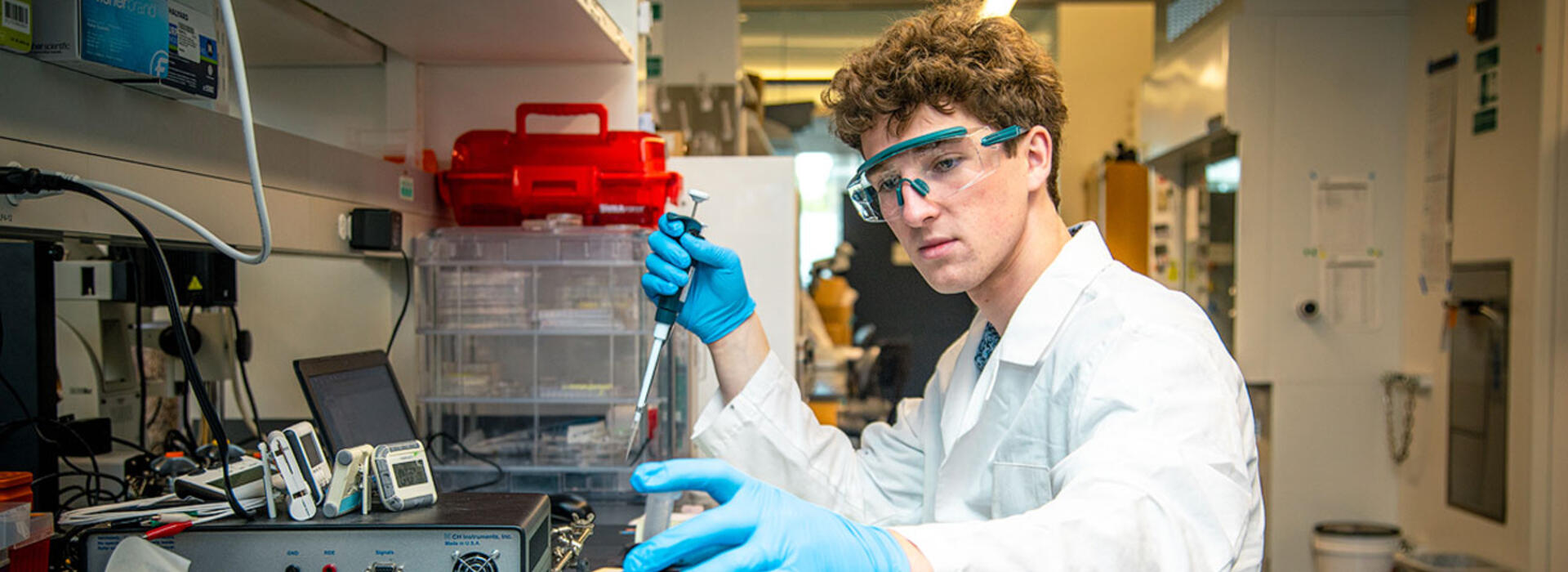 A man in a white lab coat sits at a lab bench, handling equipment and taking notes.