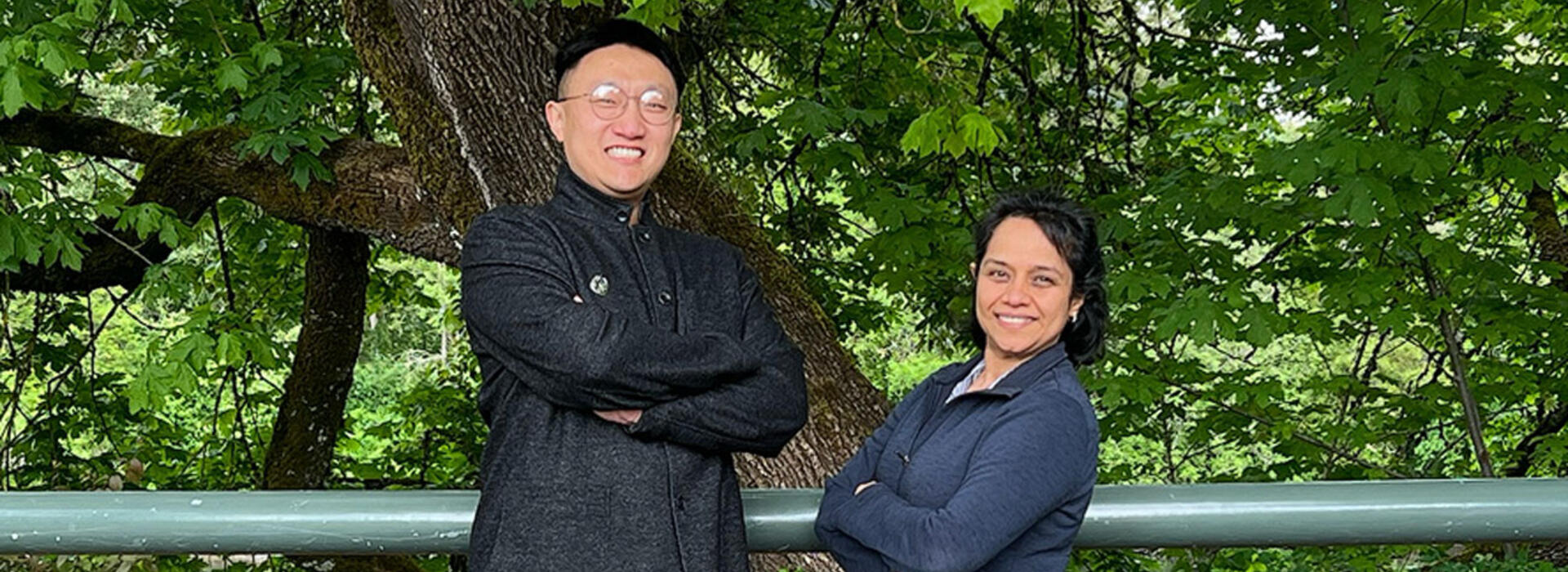 Two people wear black and stand outside in front of vegetation and smile toward the camera.