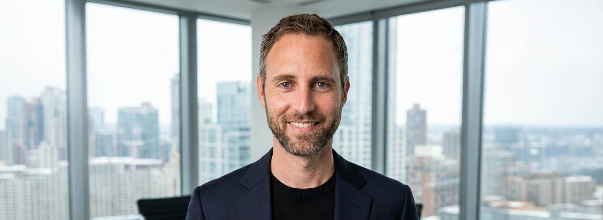 A person wears a dark suit in an office setting and looks toward the camera with tall windows and a city skyline in the background.