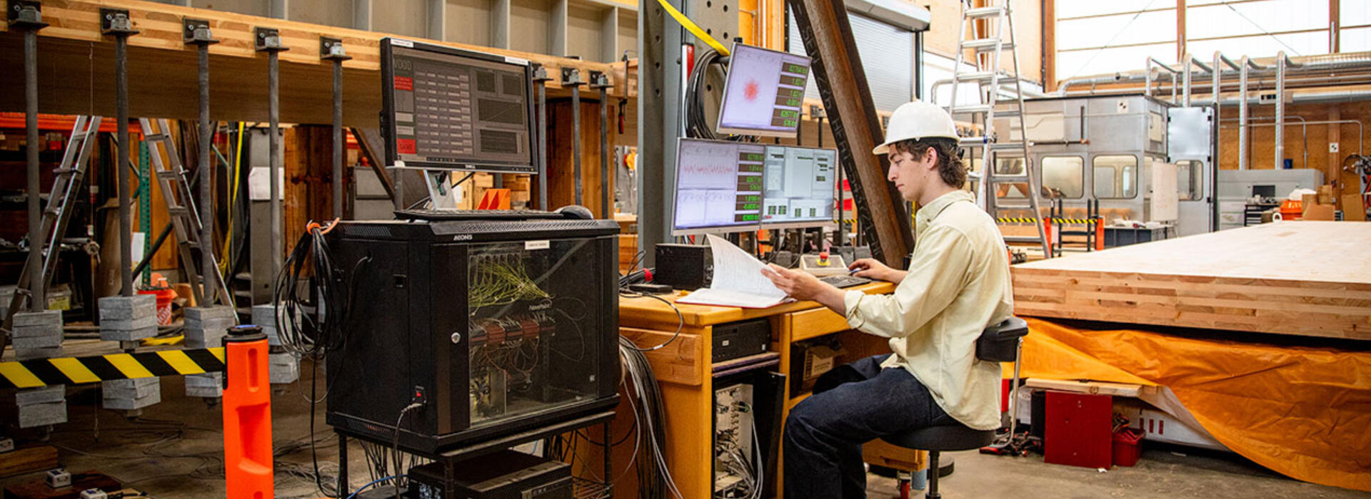 Dylan Goff wears a hard hat and sits in front of a computer and monitoring equipment in the Emmerson Lab, a large timber engineering facility.