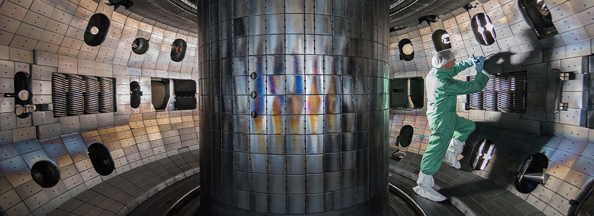 A person wears a clean-room suit with hair covering, gloves, and boot covers and stands inside a large cylindrical chamber lined with metallic panels and equipment.