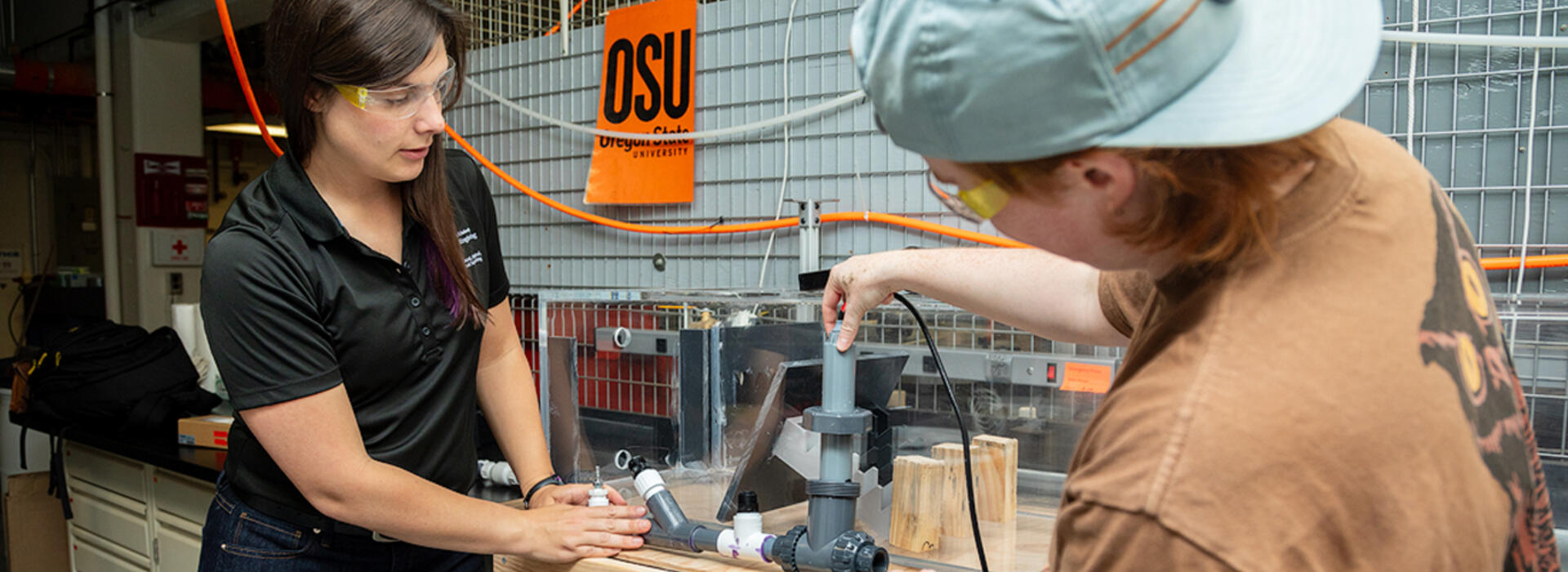 Kathryn Newhart and Perry Wilson wear eye protection and stand next to a workbench in a lab and adjust a device with two intersecting pipes.
