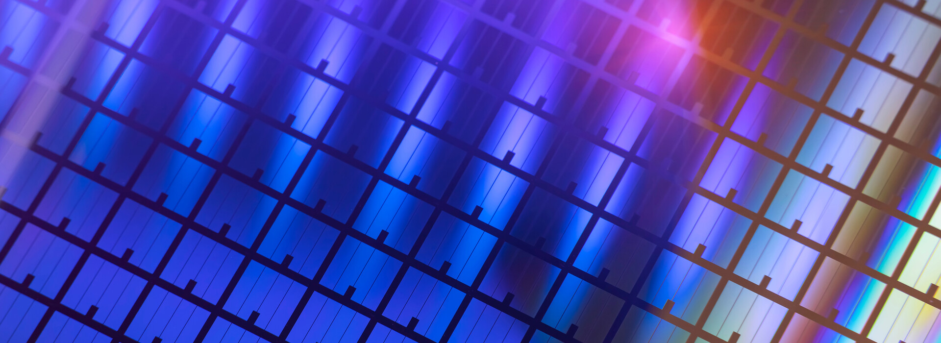 A close-up view of a semiconductor wafer with a repeating grid pattern of microelectronic structures in iridescent colors.