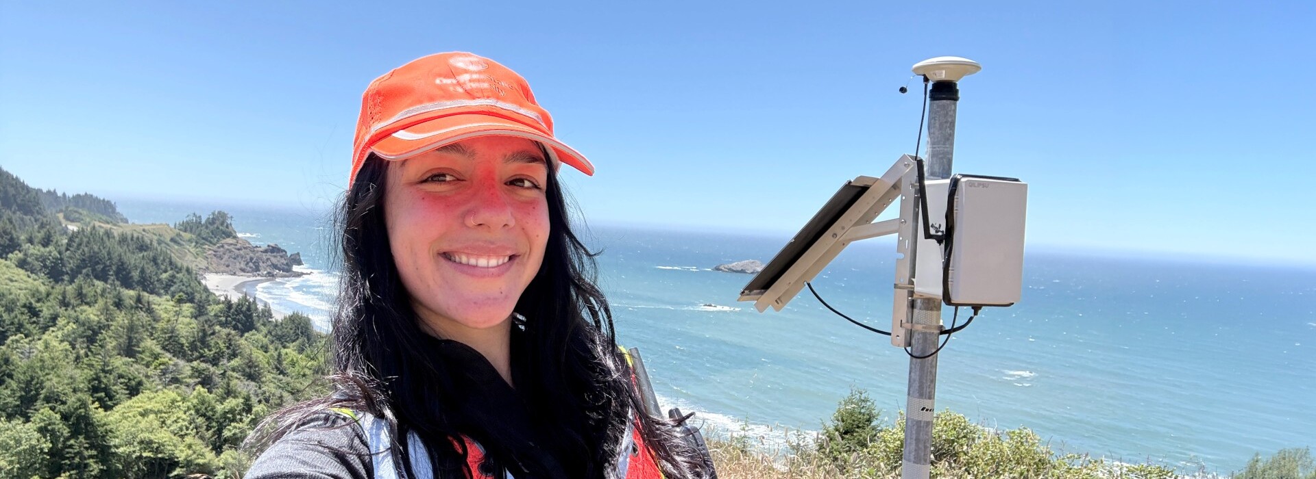 Heather Maran stands beside a coastal monitoring instrument overlooking the Oregon Coast with vegetation and ocean behind her.