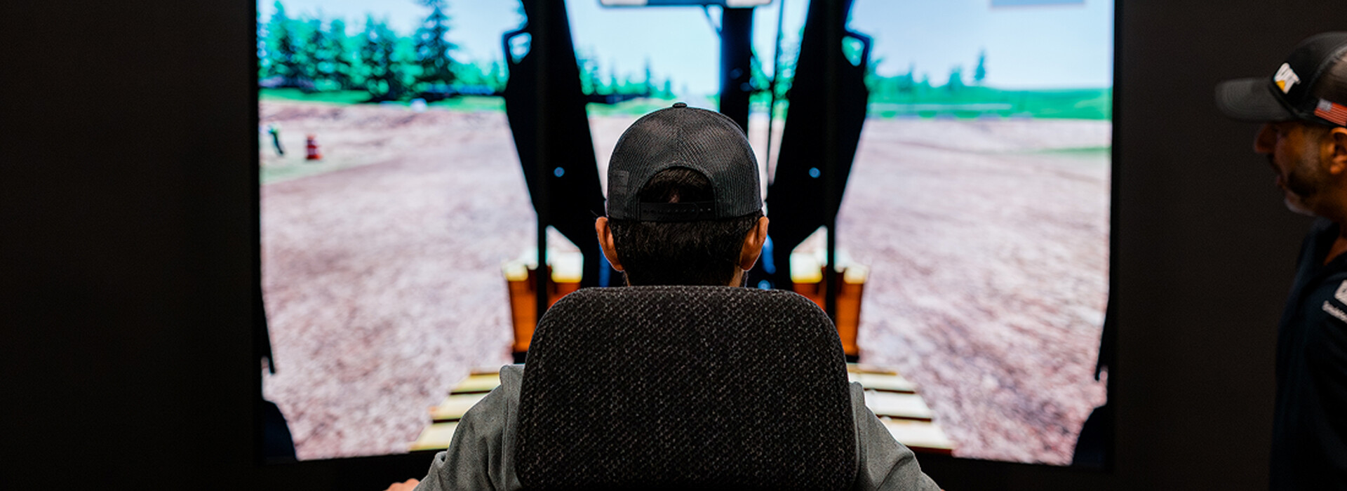 A rear view of a person seated in a heavy equipment simulator, facing a large screen that displays a virtual construction site.