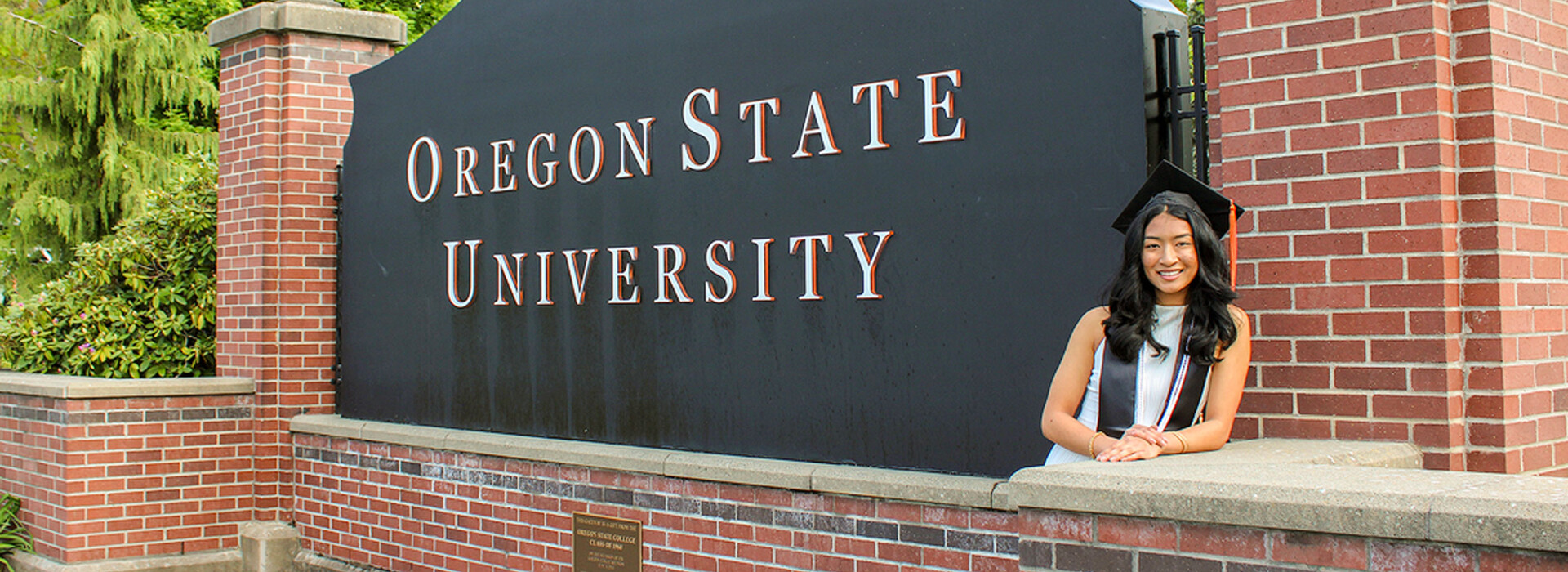 Megan Thomas wears a graduation cap and stole and stands outdoors leaning against a large black Oregon State University sign.
