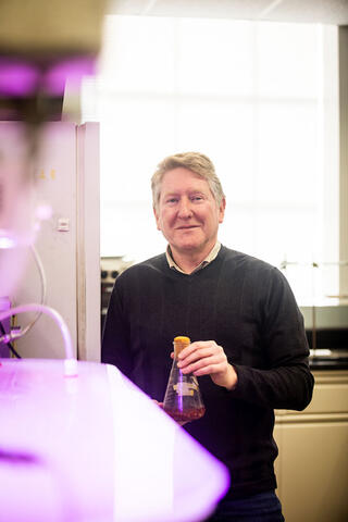 A person stands in a lab and holds a glass flask with a dark liquid looking toward the camera.