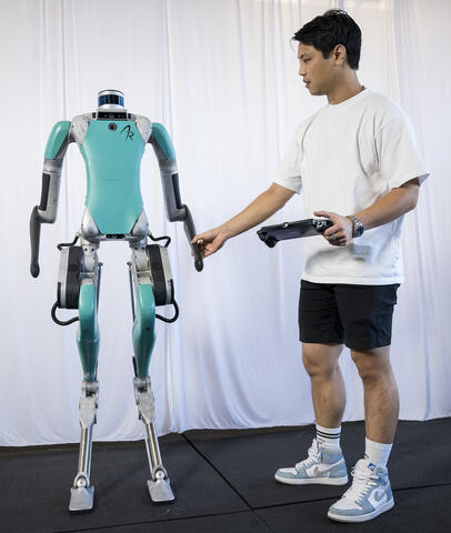 Researcher standing beside a humanoid robot with a controller in hand.