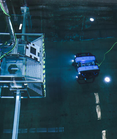 An underwater testing facility with a metallic structure and an unmanned underwater vehicle with lights, connected by a green tether.