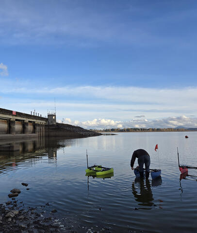 A person with three small robotic research boats near a dam structure on a lake under a blue sky.