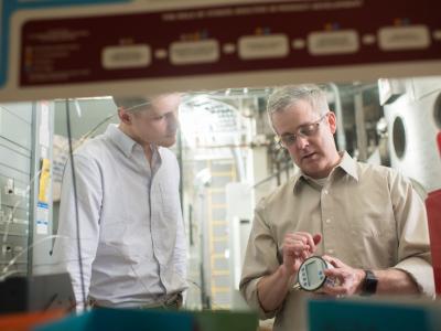 Two faculty members watching a circular piece of machinery. The person on the right holds the piece of equipment and points to it. 