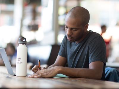 A person writing on a table with his laptop and water bottle in front of him. 
