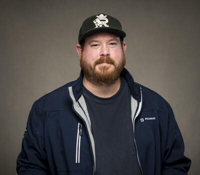 Bearded man in a dark jacket and cap stands against a neutral background. His expression is neutral.