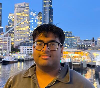 A person looking at the camera, wearing glass. High rise buildings in the background.
