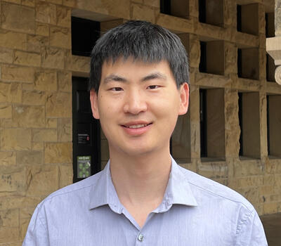 A person in a light blue shirt smiles in front of a stone building with square windows.